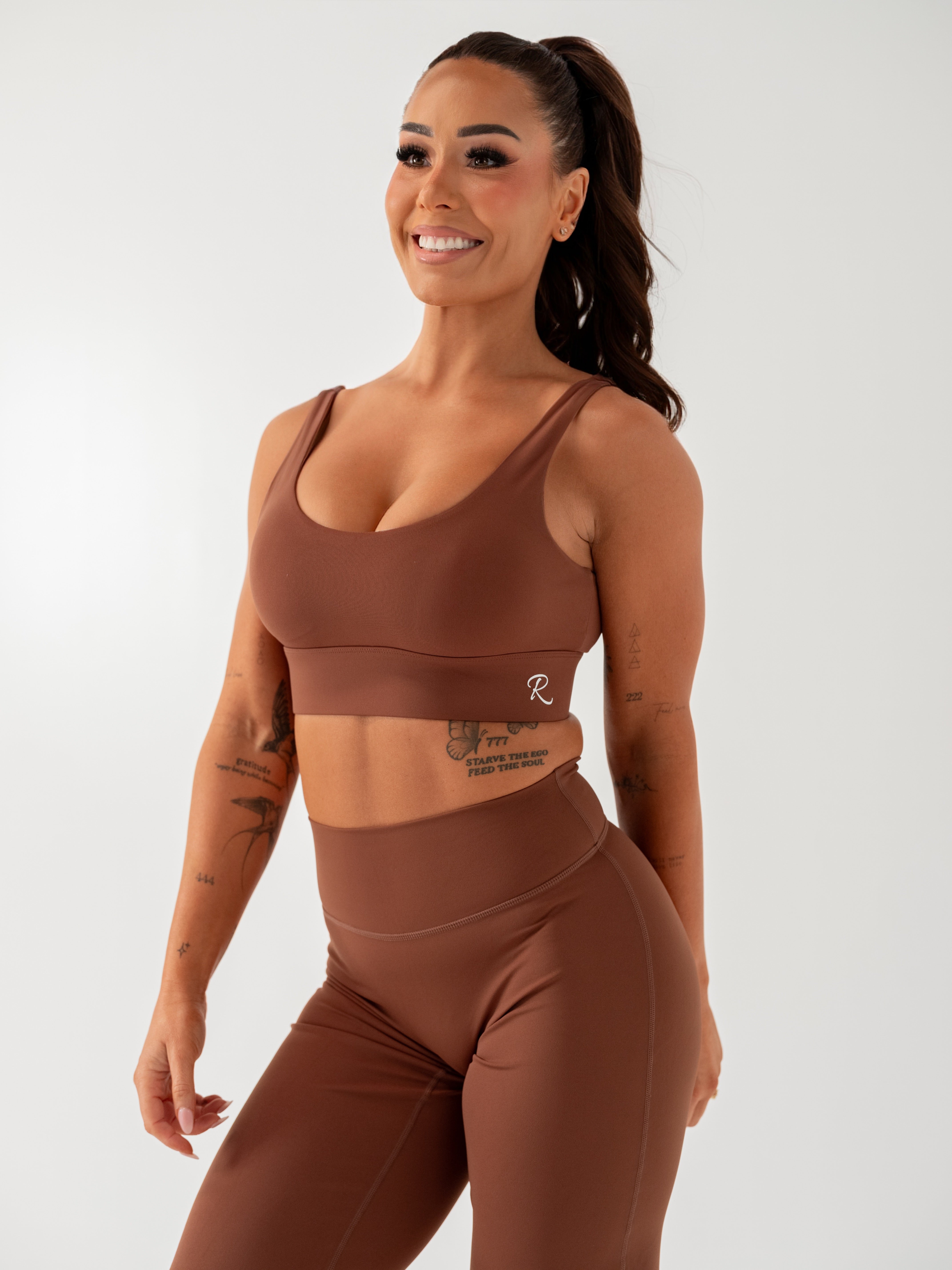 A woman modeling a matching brown activewear set consisting of a scoop-neck sports bra and high-waisted leggings, photographed against a clean light gray studio background.