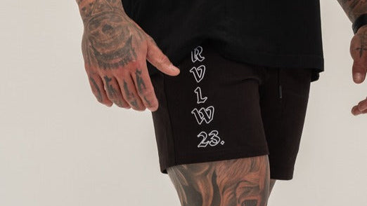 Person wearing black shorts with white text and tattoos on arms and legs against a plain background